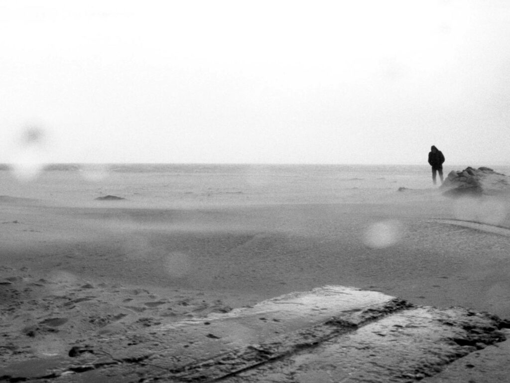 Schwarz-weiß Foto von einem Spaziergänger am Strand bei schlechtem Wetter